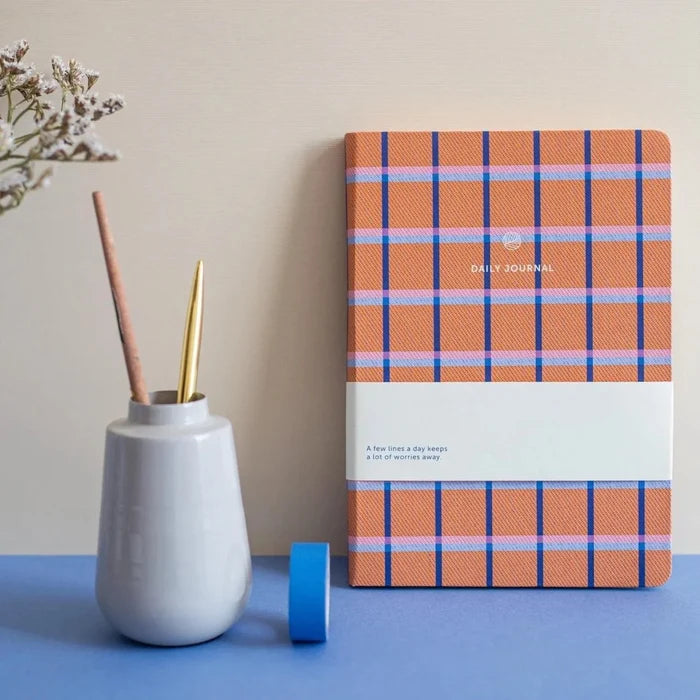 Plaid A-Journal notebook with 'Daily Journal' on a blue surface next to a white vase with a gold pen.