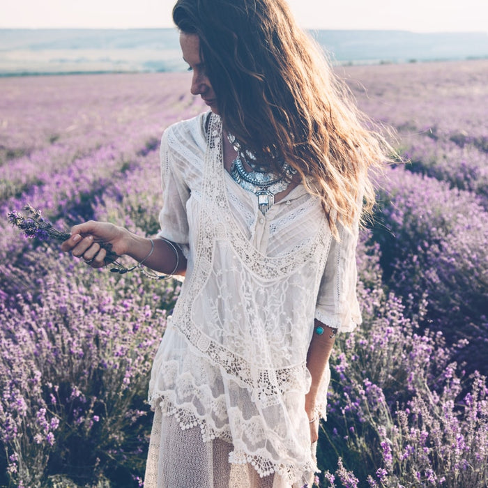 A woman in a white lace dress standing in a lavender field holding a small bouquet of lavender, evoking calm and natural wellness for Bath & Body Oil | Privai.
