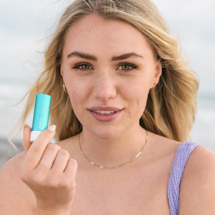 Young woman with natural makeup holding Classic Liplux® Organic Lip Balm Sunscreen SPF 30 - Original | COOLA near her face, promoting skincare and sun protection outdoors.