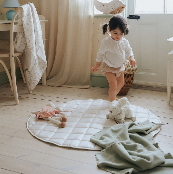 Child playing on a quilted mat with toys in a room with wooden floor and white walls, with a green blanket in the foreground.