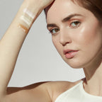 Close-up of a woman showing arm swatches of three shimmering highlighter shades labeled Cosmos, Solstice, and Eclipse using Glow Time Highlighter Stick | Jane Iredale, highlighting skincare and beauty wellness.