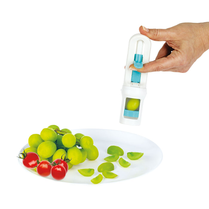 A hand uses the Nuby Grape Cutter | Grey to slice a green grape over a white plate with sliced grapes, whole grapes, and cherry tomatoes, making safe baby food prep easy.