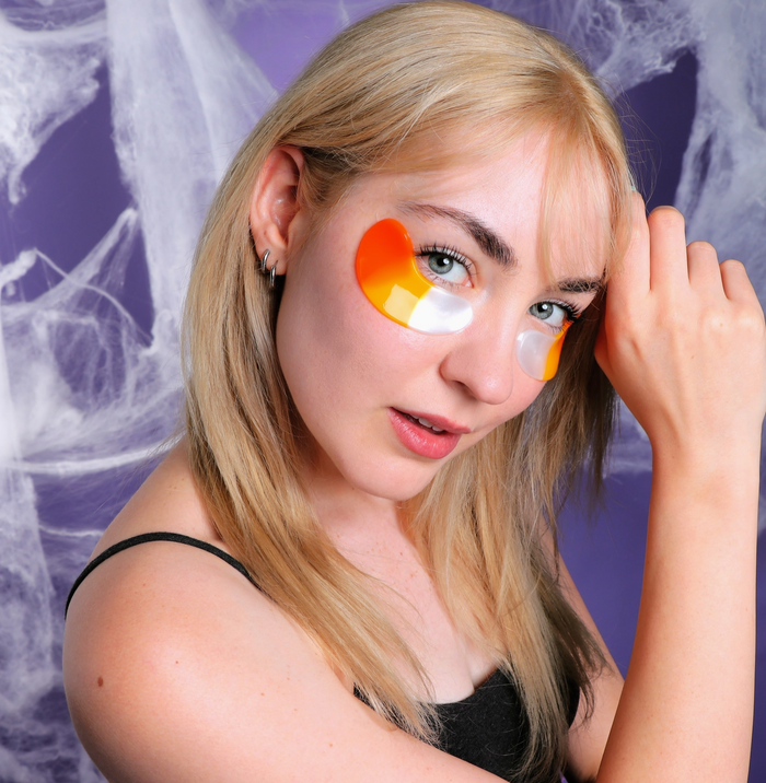 A blonde woman wearing orange and white crescent-shaped eye patches, posing against a purple background with white spiderwebs.
