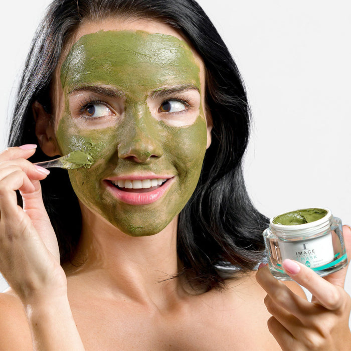 Woman applying a green facial mask from a jar, showcasing skincare and wellness with I MASK purifying probiotic mask | IMAGE Skincare for a refreshing spa experience.