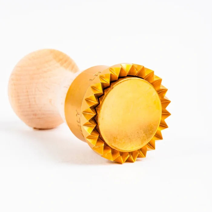 A wooden handle pasta stamp with brass components, featuring apparent flaws as part of its artisan production on its side on a white background.