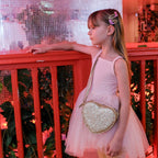 Young girl in a pink dress holding a heart-shaped glittery purse against a red railing.