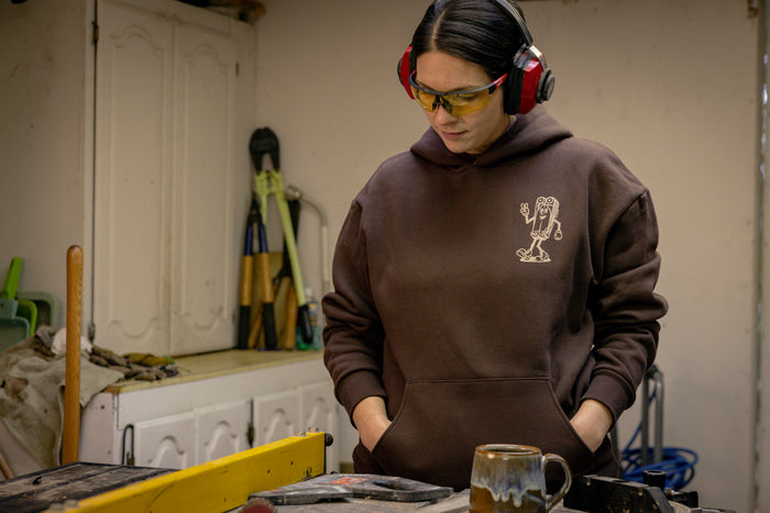 A person wearing a brown Leatherman Mr. Wave hoodie standing in a workshop with safety glasses and ear protection, looking down at a workbench with tools and a ceramic mug.