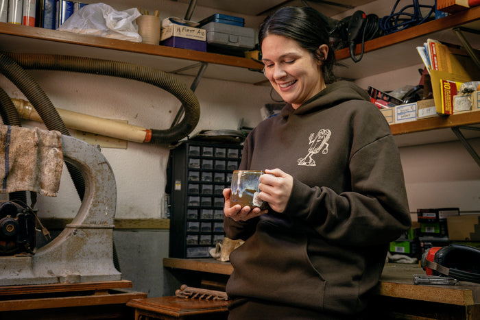 A person wearing a brown Leatherman Mr. Wave hoodie standing in a workshop holding a ceramic mug, smiling while surrounded by tools and equipment.