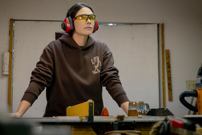 A person wearing a brown Leatherman Mr. Wave hoodie standing at a workbench in a workshop, wearing safety glasses and earmuffs.