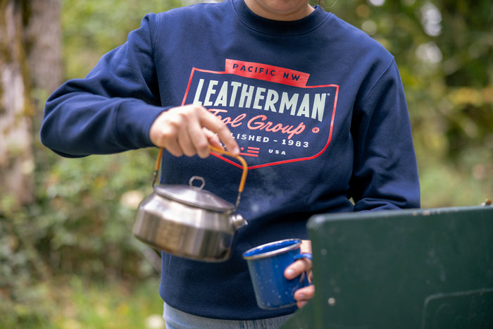 Close-up of a person wearing a navy blue Leatherman PNW Crewneck sweatshirt while pouring hot water from a kettle into a blue enamel mug at a campsite.