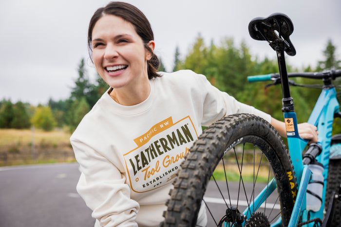 A person wearing a cream-colored Leatherman PNW Crewneck smiling while holding onto a blue mountain bike outdoors.