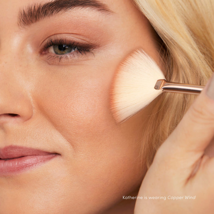 Close-up of a woman applying Purepressed Blush | Jane Iredale with a fan brush, highlighting a natural, radiant complexion and subtle warm tones.