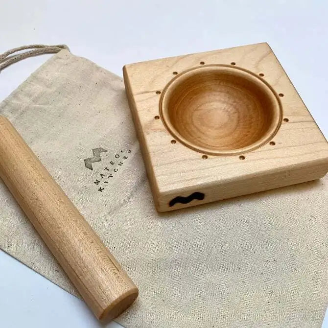 A square maple wood ravioli mold with a circular indentation in the center, with carry bag and rolling pin on white surface.