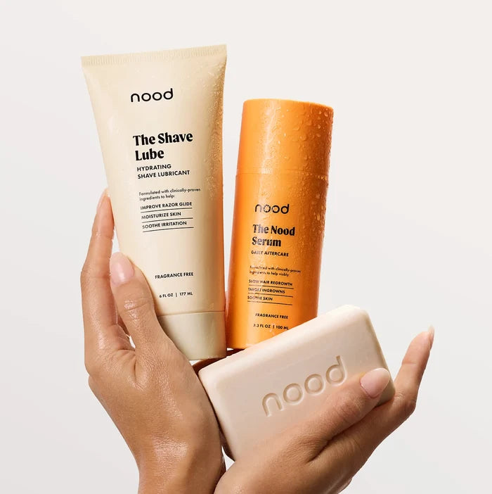 Hands holding Nood Shave Collection including The Shave Lube hydrating shave lubricant, Nood Serum daily aftercare, and Buff Bar pre-shave soap on light background